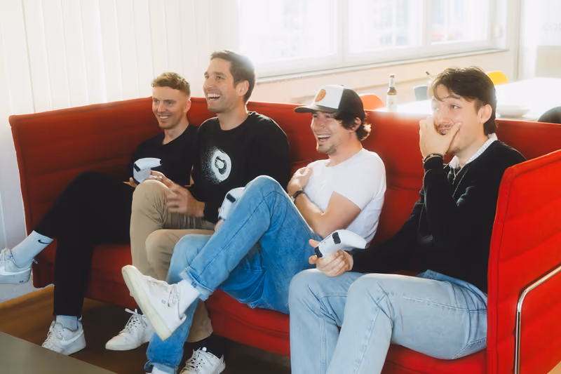 Vier Kollegen sitzen lachend mit Controllern in der Hand auf dem roten Sofa im Büro