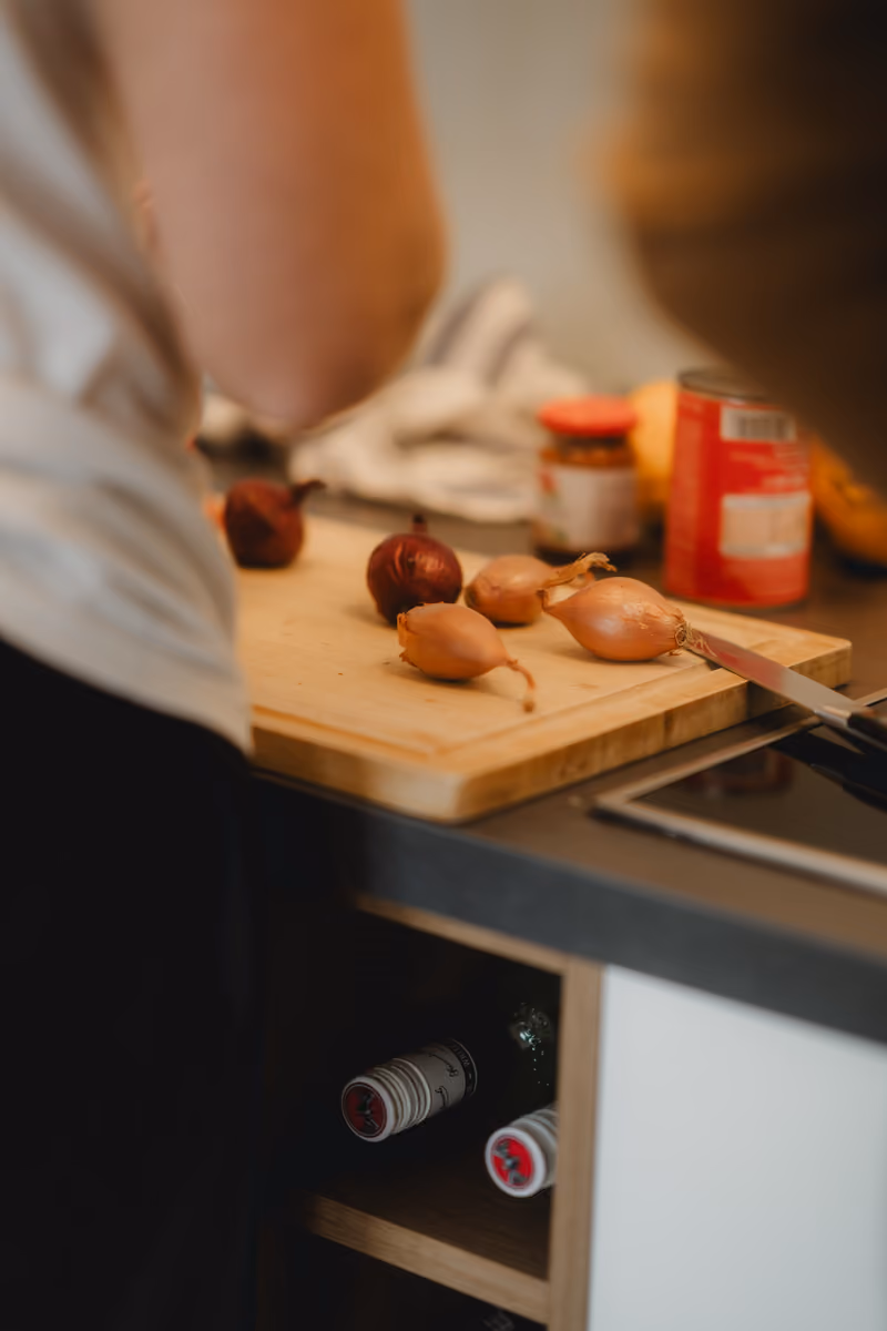 Nahaufnahme eines Schneidebretts mit Zwiebeln und Schalotten in der Büroküche, dahinter jemand beim Vorbereiten
